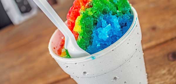 brightly colored shave ice in white cup with spoon on wooden table